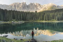 A person in a red jacket stands on a rock, gazing at a serene lake. The water reflects a stunning image of distant mountains and a dense forest under a clear sky, creating a mirror-like effect. The scene exudes tranquility and natural beauty.