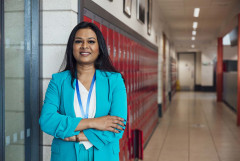 An educator in a turquoise suit stands in a school hallway in front of a row of red lockers.