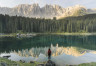 Person standing on rock at the edge of a reflective lake with a forest and mountains in the distance.