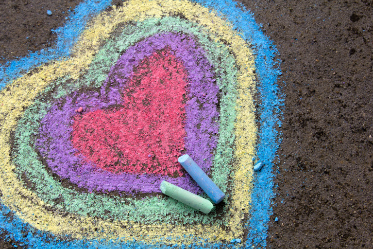 A colorful heart is drawn on the pavement with chalk, featuring layers of red, purple, green, yellow, and blue, with two pieces of chalk lying nearby.