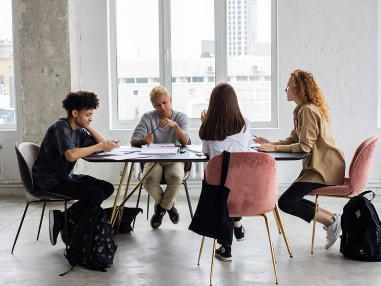 Four young adults are seated around a table in a bright, modern room with large windows, engaged in discussion or collaborative work, with backpacks on the floor and papers spread out on the table.