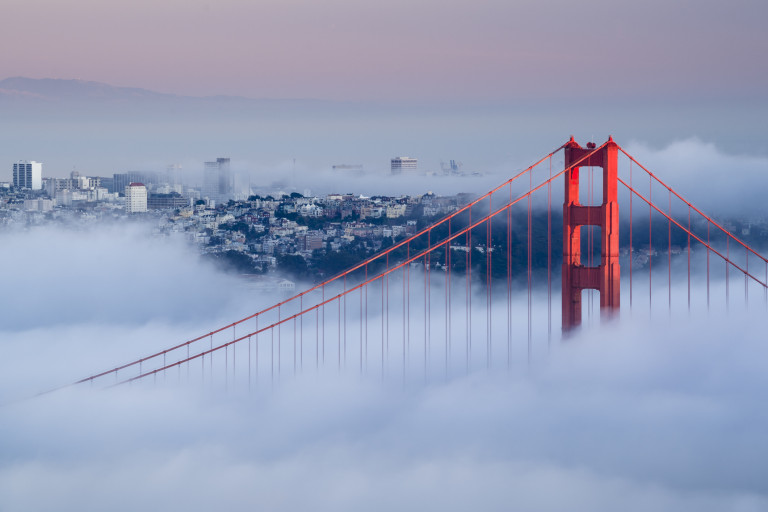 The top of the Golden Gate Bridge peeks through a thick blanket of fog, with the cityscape of San Francisco in the background, bathed in the soft light of early morning or late evening.