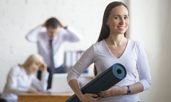 A woman walking out of her office away from her colleagues with a yoga mat on her hand and smiling to the camera