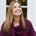 an image of Threlkel Leigh smiling with long blonde hair, wearing a dark red top and a necklace with round pendants