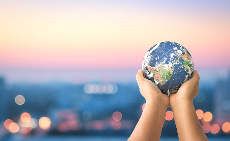 Two hands hold a small globe against a backdrop of a softly blurred cityscape at dawn or dusk. The sky shows gradients of pink, orange, and blue, with distant lights creating a bokeh effect, resembling stars in the background.