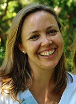 A smiling woman with light brown hair stands outdoors, wearing a light-colored top